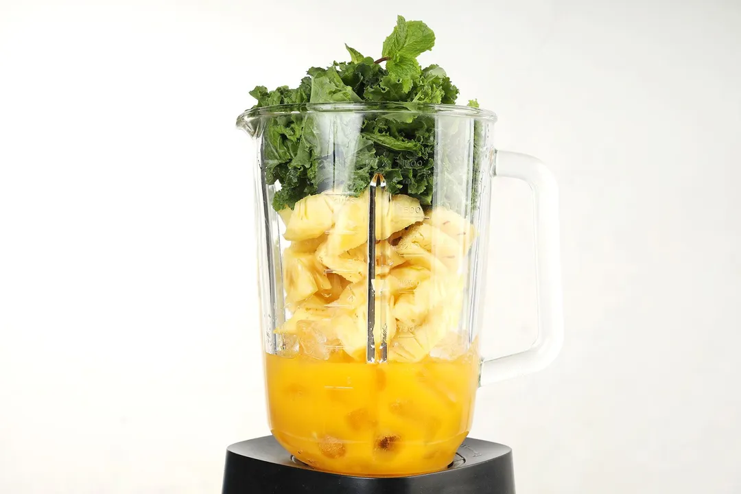 A blender filled with orange juice, pineapple slices, ice, and kale.