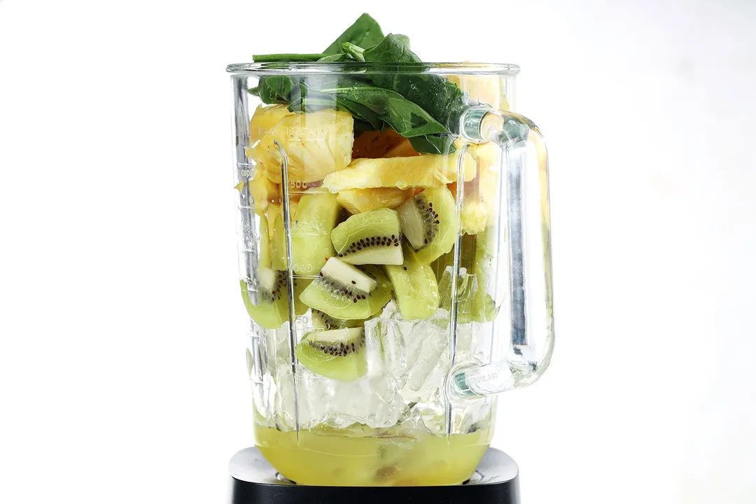 A blender filled with ice, green kiwi slices, pineapple slices, and spinach.