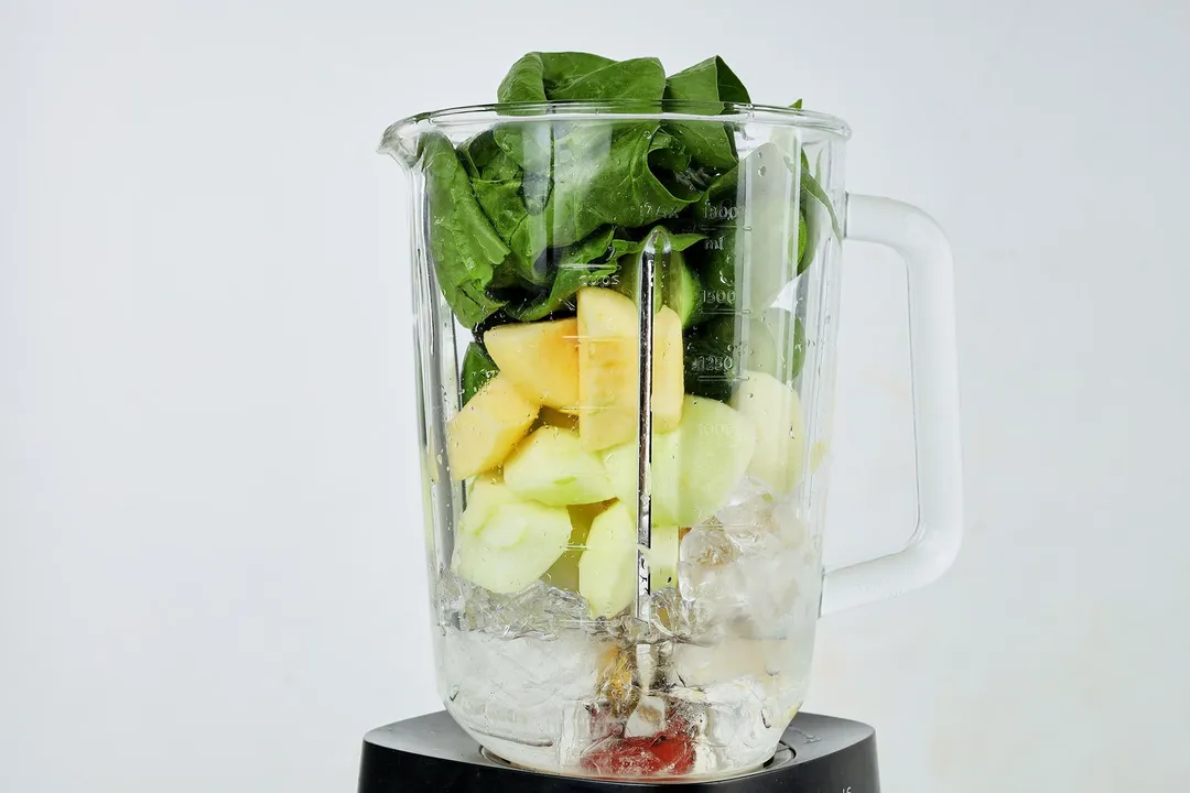 A blender filled with layers of crushed ice, apple wedges, and spinach leaves.