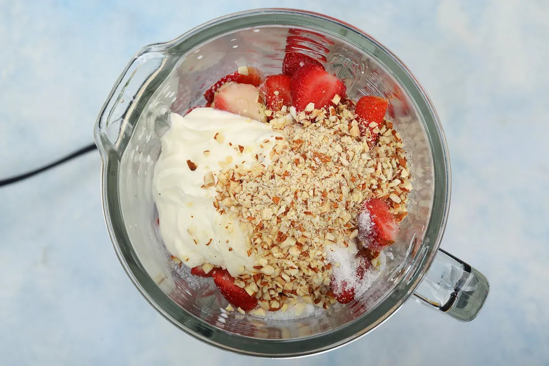 A high-angle view of a blender filled with strawberries, yogurt, and chopped almond.