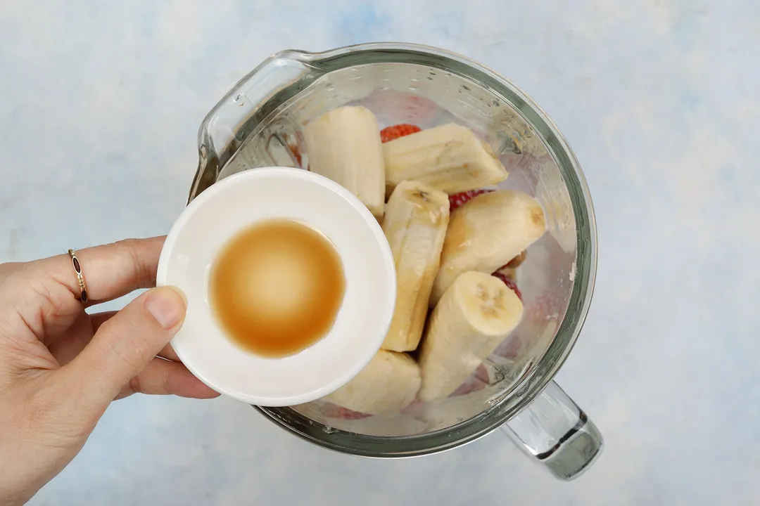 A hand holding a small plate with some honey in the center over a blender filled with sliced bananas.
