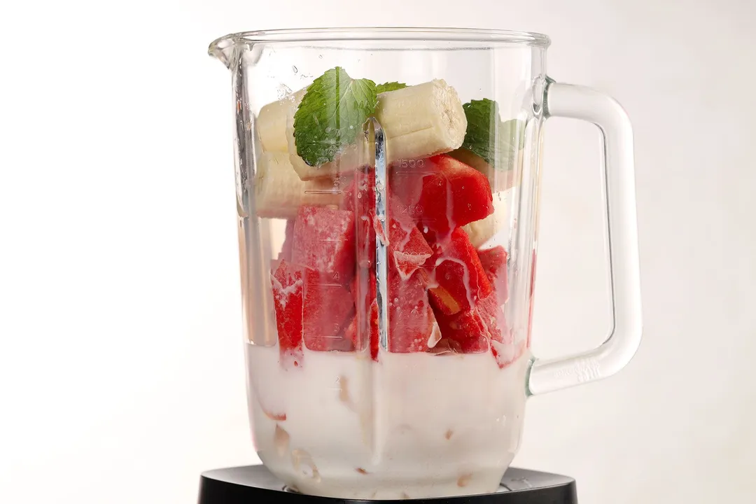 A blender filled with ice, watermelon slices, peeled bananas, and mint leaves.
