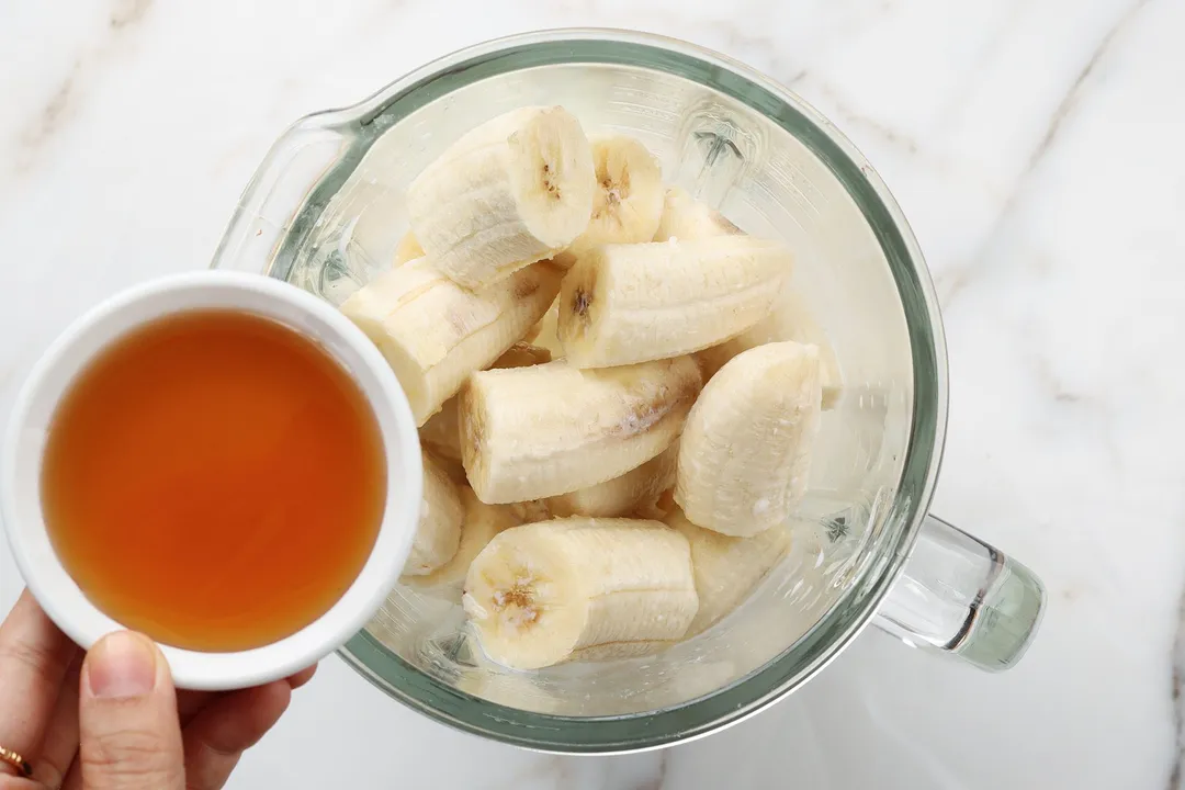 A high angle shot of a blender filled with sliced banana and a hand hovering above it holding a small plate of honey.