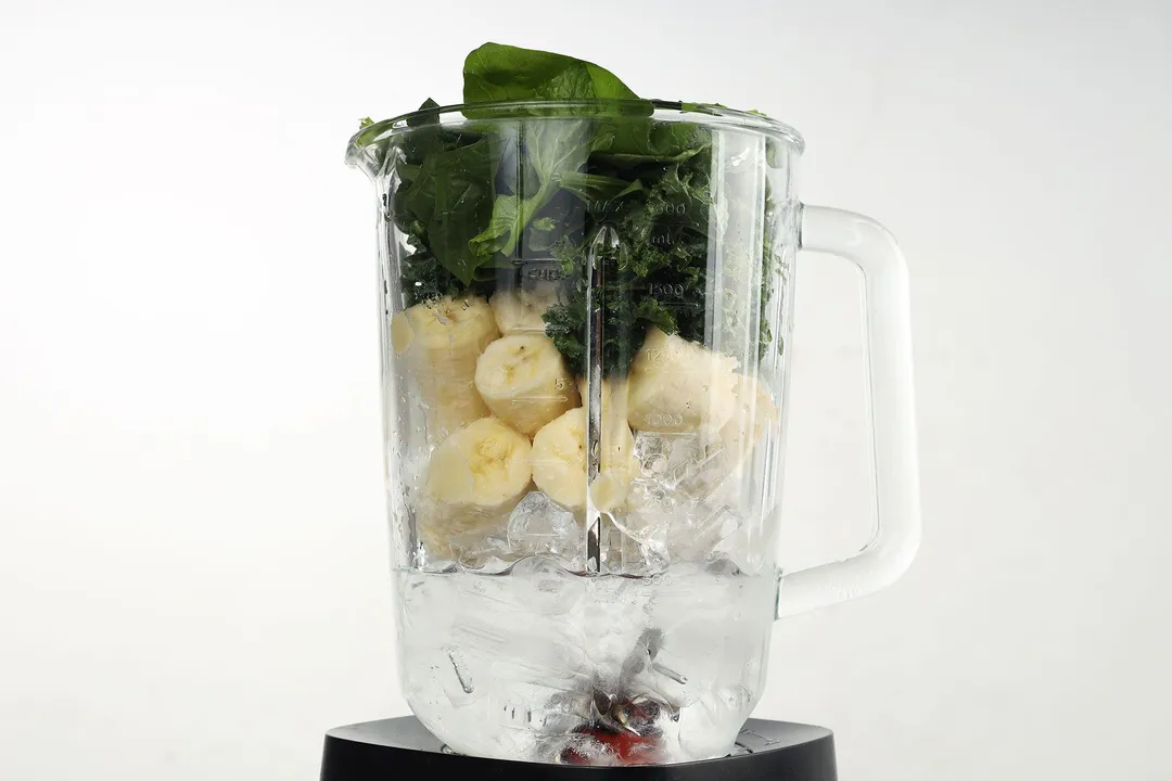 front shot of a blender pitcher with kale, spinach, banana cubed and ice in it