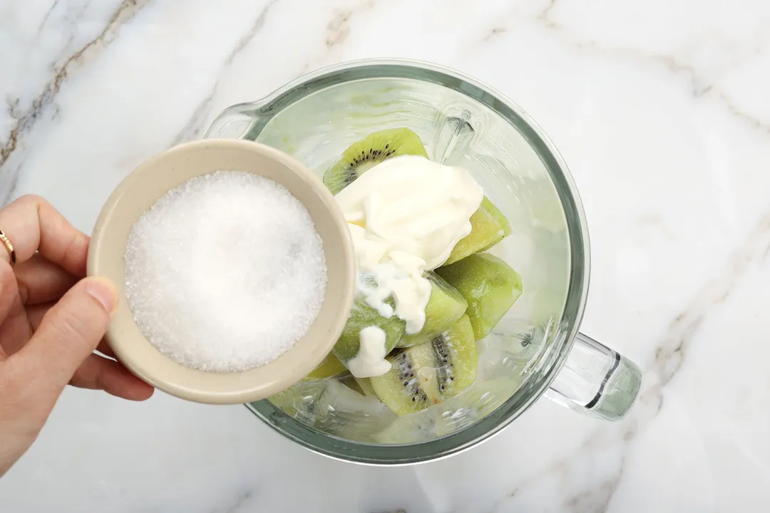 a hand holding a small bowl of sugar on top of a blender pitcher with kiwi sliced and yogurt in it