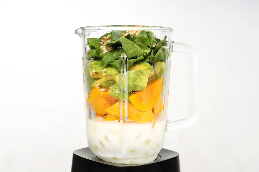 front shot of a blender pitcher with milk, mango cubed, avocado cubed, spinach and almond in it.