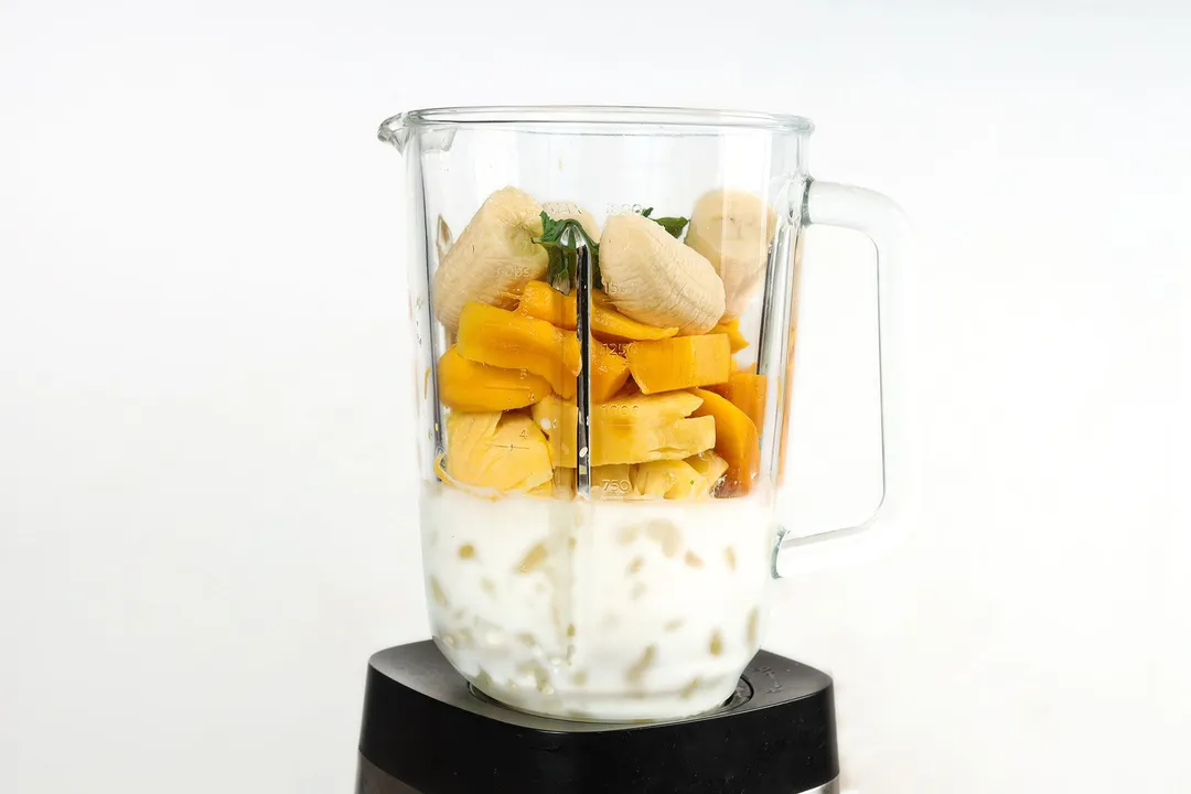 a front shot of a blender pitcher with banana, mango cubed, pineapple cubes, milk and ice in it.