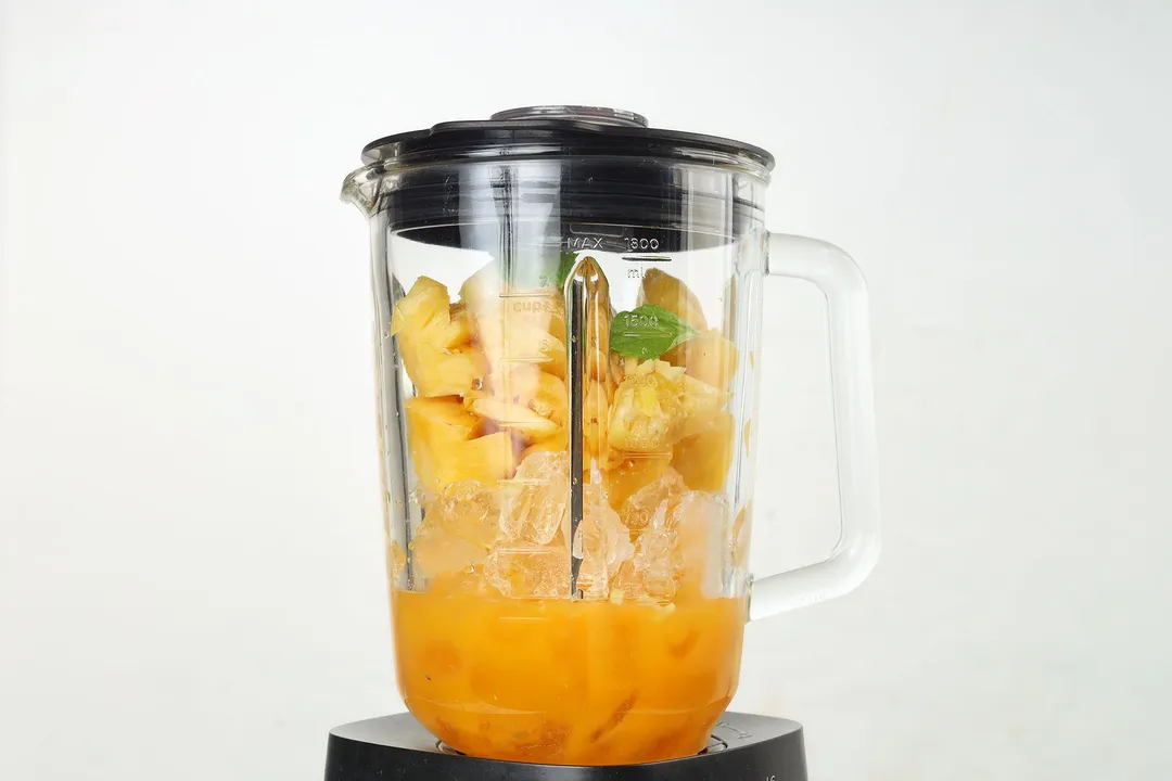 a blender of pineapple cubed, mango cubed, ice, orange juice and mint leaves