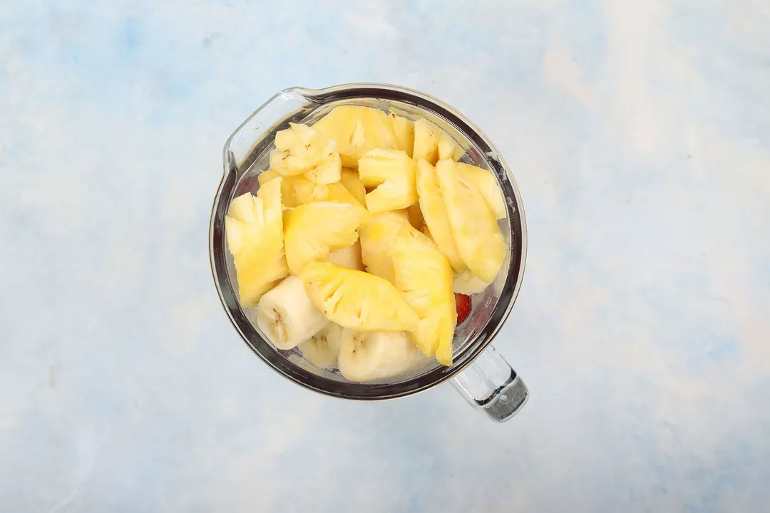top view of a blender pitcher full of pineapple cubes, banana cubes and strawberry