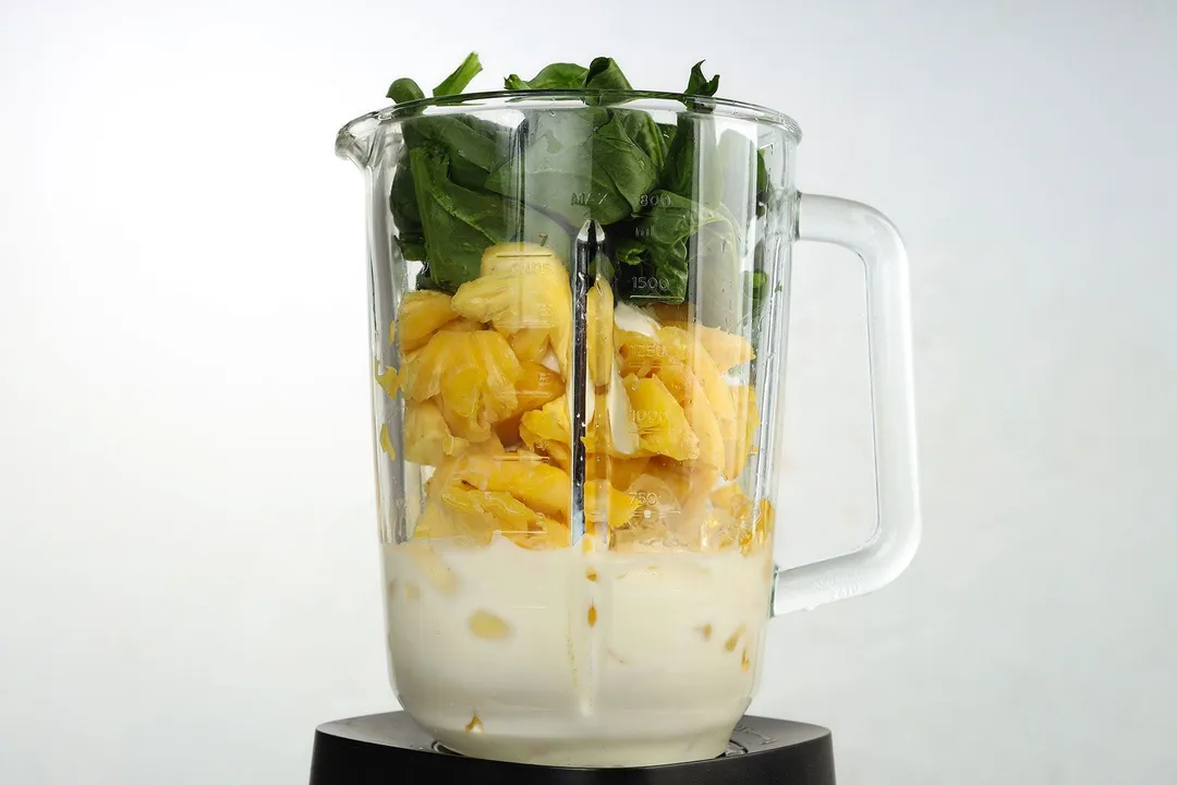 font shot of a blender pitcher full of spinach, pineapple cubed, milk and ice