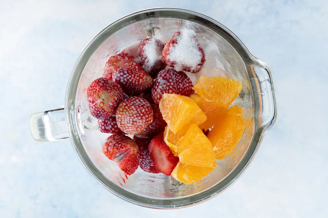 a blender pitcher full of sliced orange, strawberries and surgar