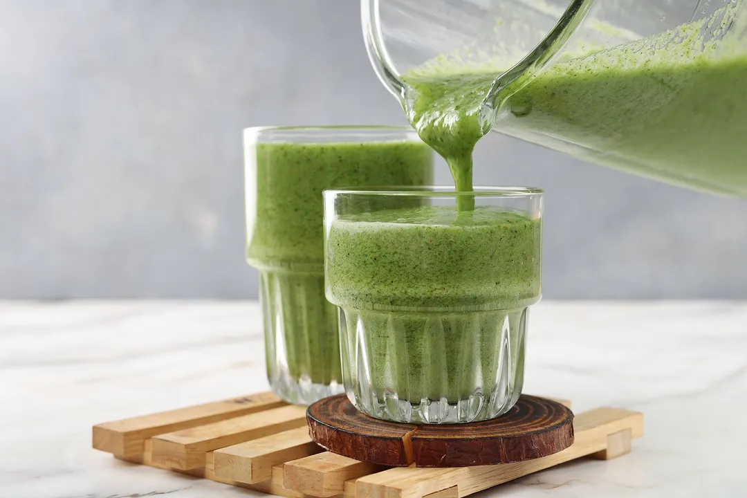 A blender pouring a green apple kiwi kale smoothie into two glasses placed on wooden coasters.