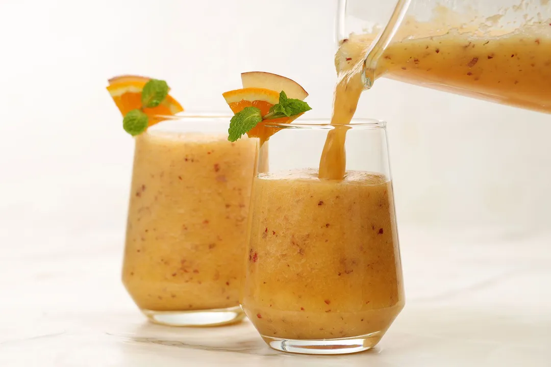 A jug pouring apple orange smoothie into a glass garnished with slices of apple and orange. A similar glass is in the blurred background.