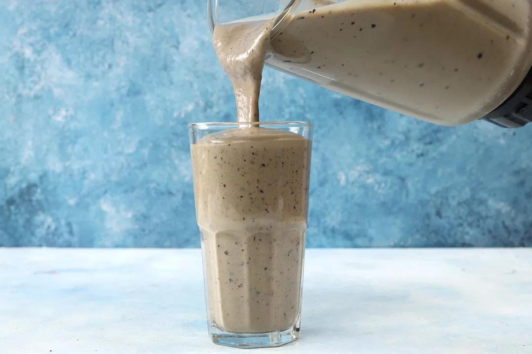 A jug pouring blueberry avocado smoothie into a tall glass.