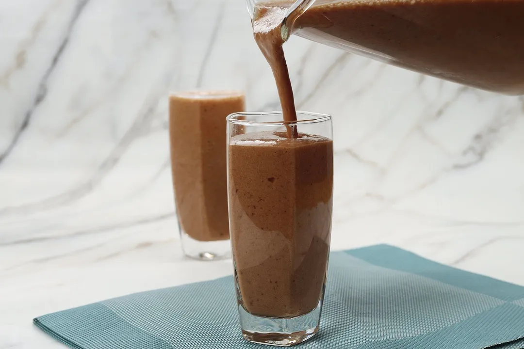 A blender pouring chocolate banana smoothie into a tall glass. 