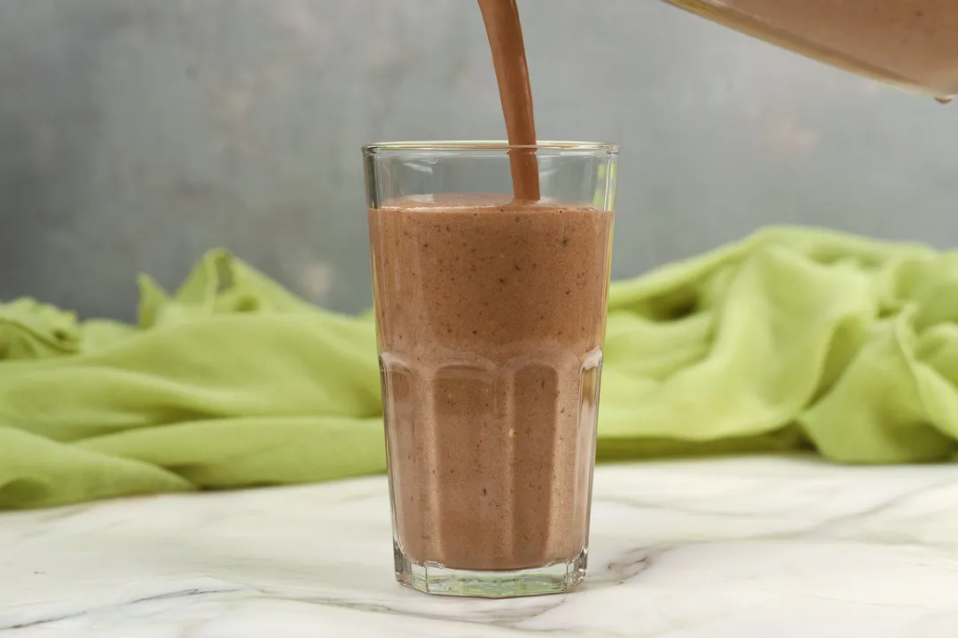 A blender pouring chocolate peanut butter banana smoothie into a glass with a green cloth in the background.
