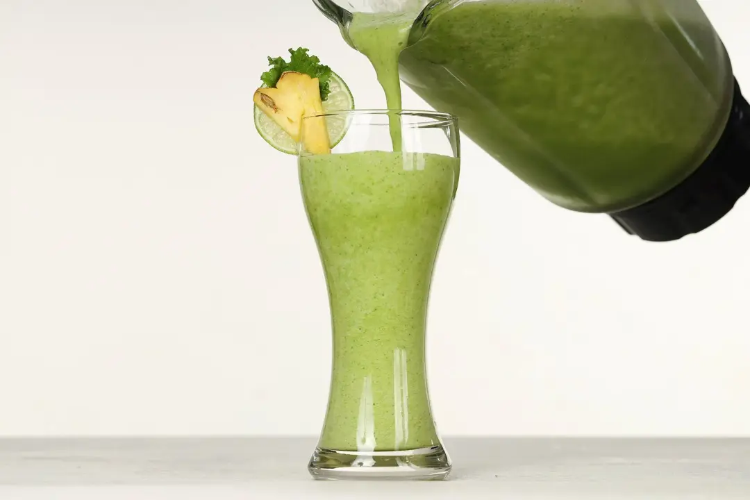 A blender of green smoothie being poured into a tall glass decorated with kale leaves and slices of pineapple and lime