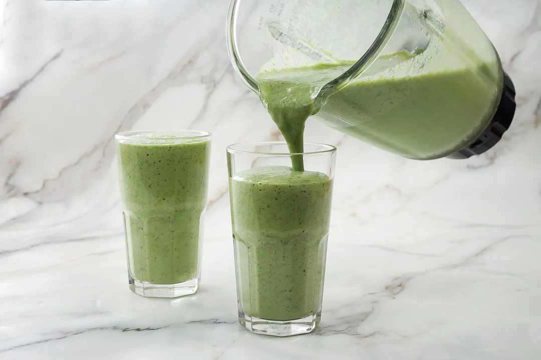 A blender pouring kiwi smoothie into two identical glasses.