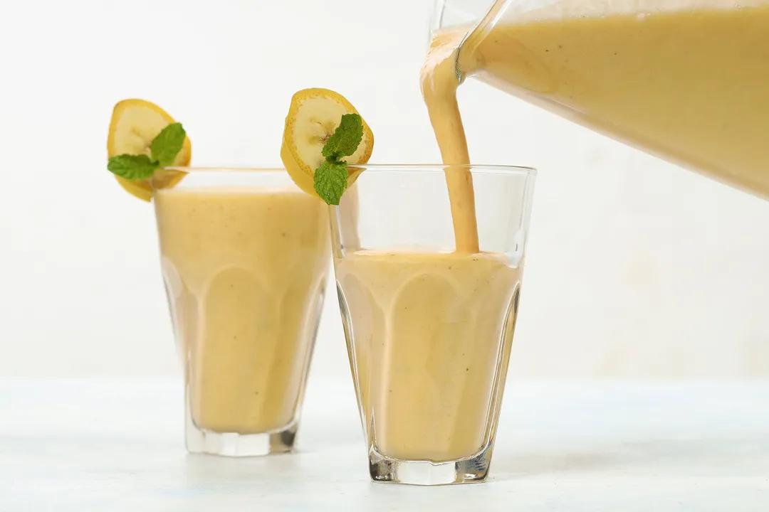 A blender pouring Mango Banana Smoothie into two identical glasses.