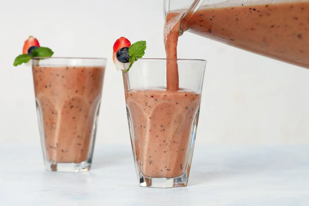 A blender pouring Mango Berry Smoothie into two identical glasses.