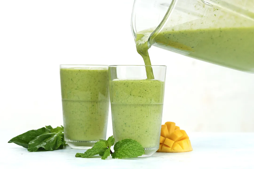 A blender pouring Mango Spinach Smoothie into two glasses.