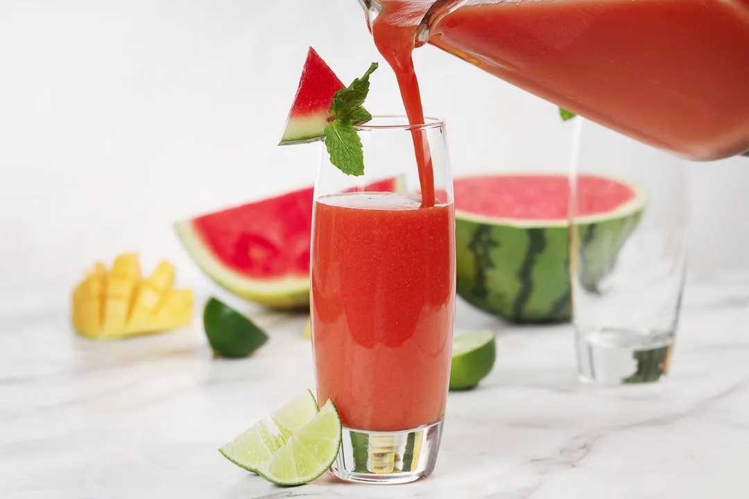 A blender pouring Mango Watermelon Smoothie into a tall glass decorated with a watermelon triangle and mint leaves.