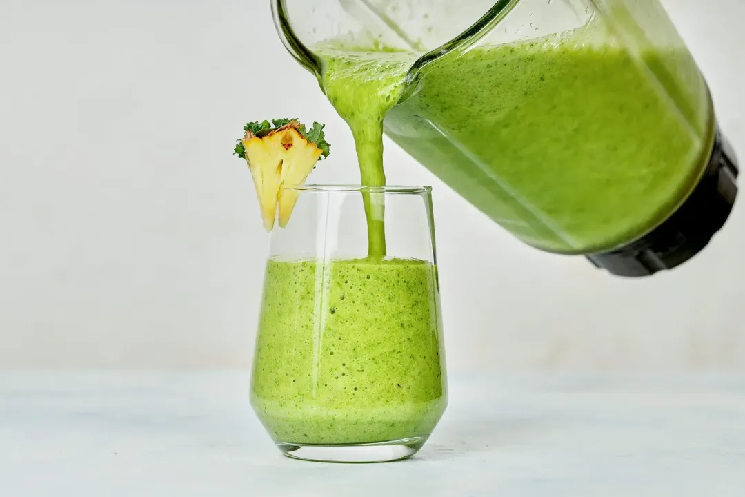 a glass of pineapple green smoothie being poured straight from the blender, with garnishes on the rim