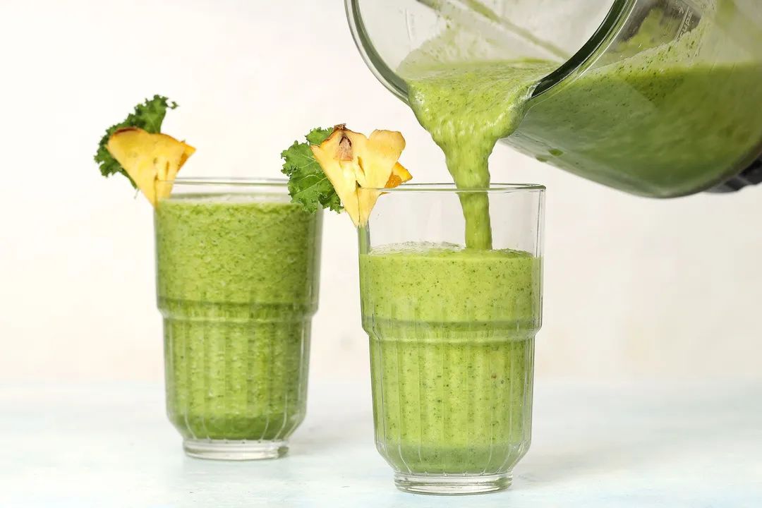 A blender pouring Pineapple Kale Smoothie into two identical glasses garnished with pineapple slices and kale.