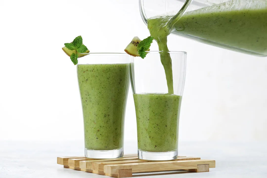 A blender pouring Pineapple Kiwi Smoothie into two identical tall glasses.