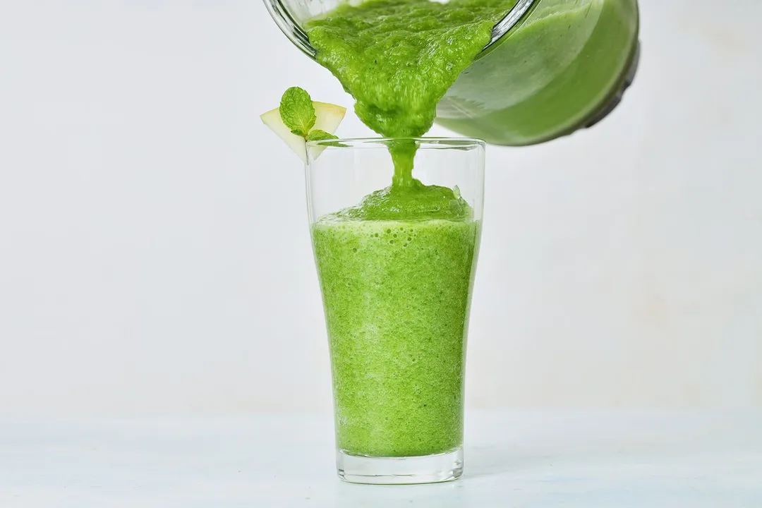 A glass jug pouring sour apple smoothie into a tall glass.