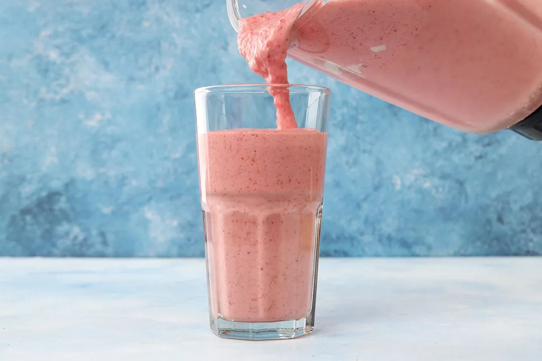 A blender pouring strawberry smoothie into a tall glass.