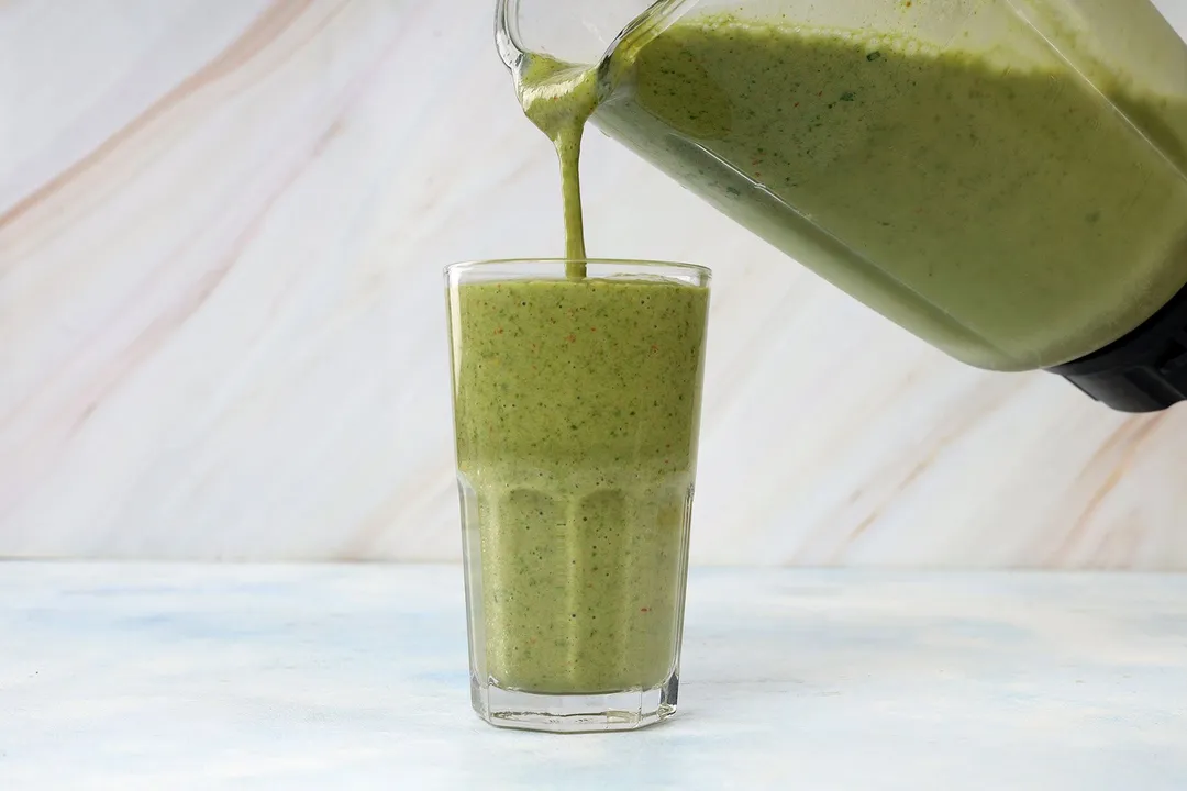 A blender pouring strawberry spinach banana smoothie into a tall glass.