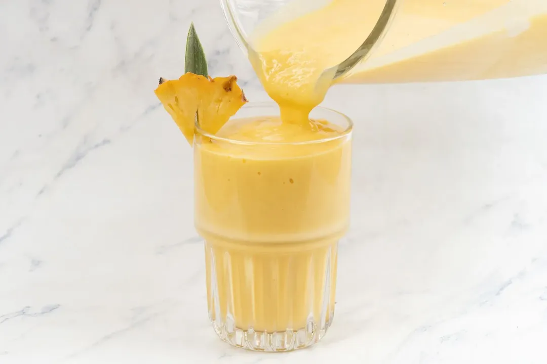 An orange-mango smoothie poured into a glass garnished with a pineapple wedge