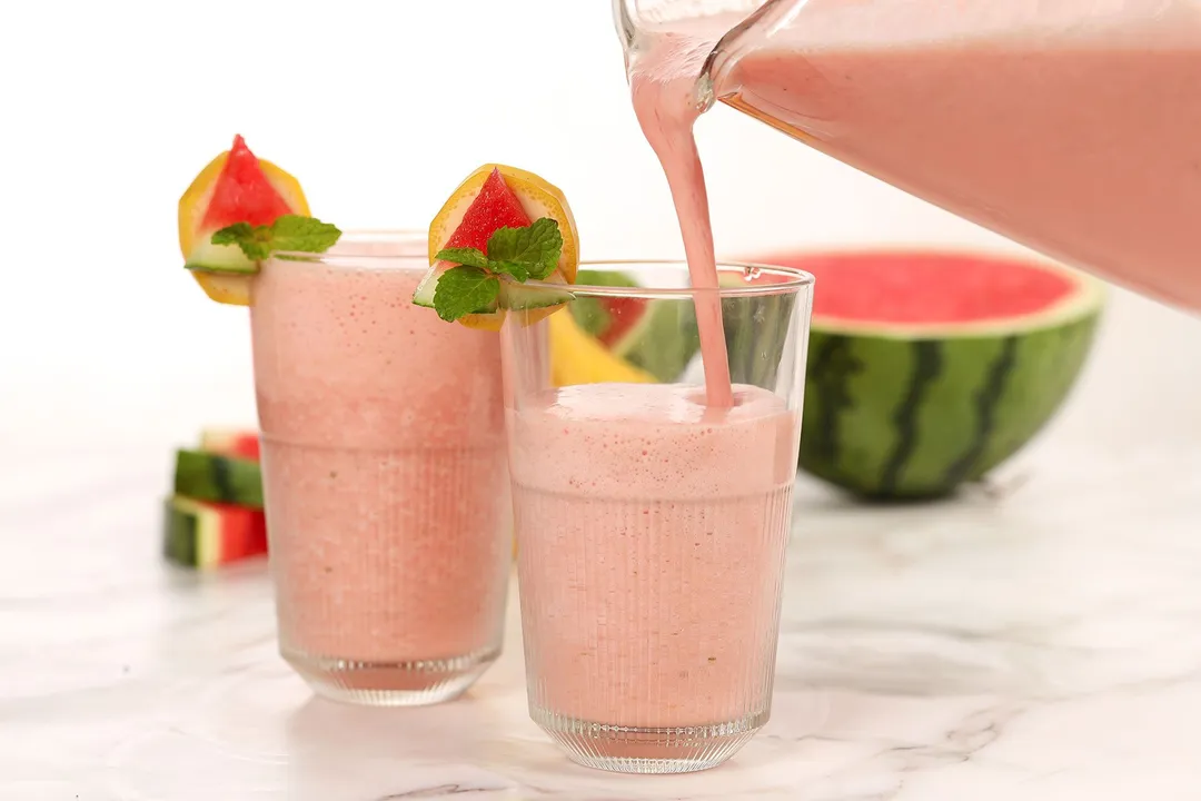 A blender pouring watermelon banana smoothie into two glasses with half a watermelon behind it.