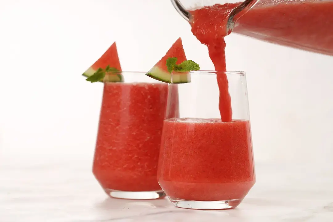 Two glasses of watermelon mojito smoothies, with one being pour from the blender 