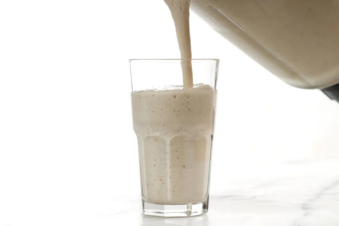 A blender pouring banana smoothie into a glass.