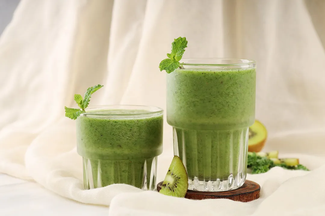 Two glasses of apple kiwi kale smoothie placed on wooden coasters on a white cloth.