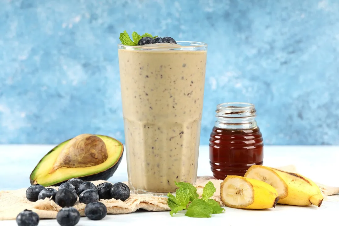 A glass of blueberry avocado smoothie placed on a wooden board and surrounded by blueberries, sliced bananas, mint leaves, half an avocado, and a jar of honey.