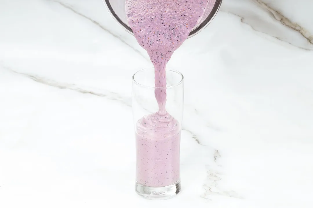 A glass jug pouring blueberry banana smoothie into a tall glass.