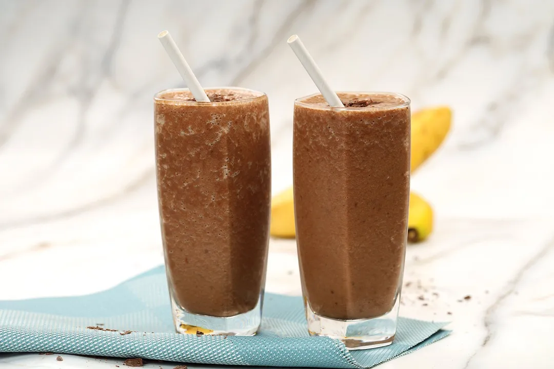 Two tall glasses of chocolate banana smoothie placed on a blue cloth.