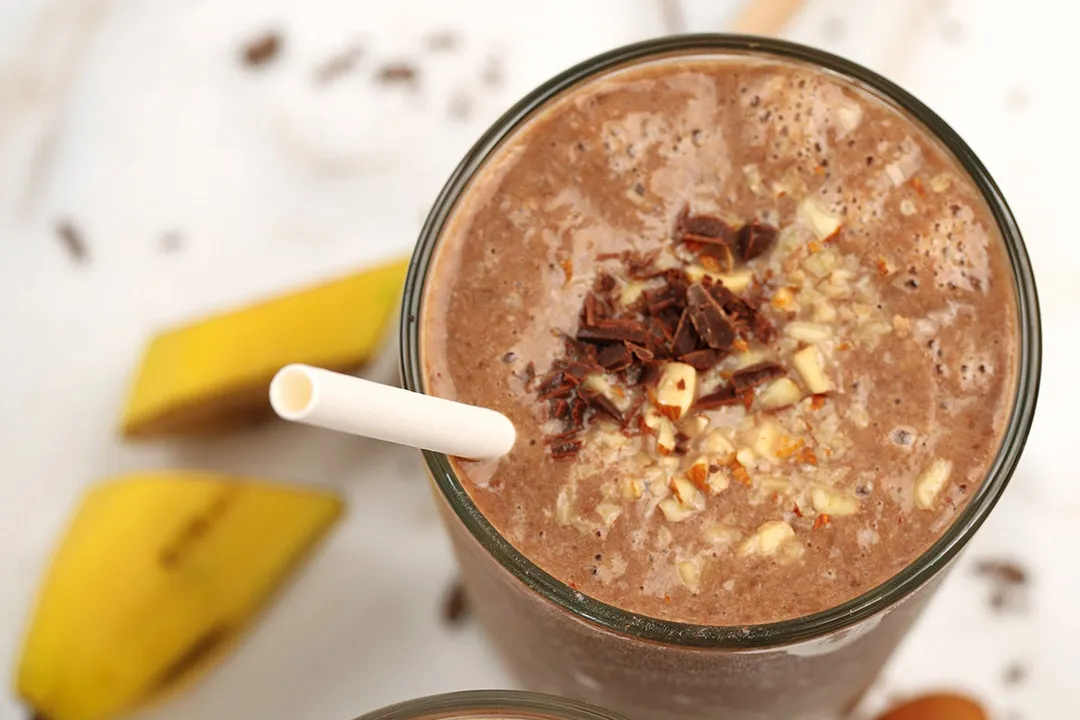 A high-angle shot of a glass of chocolate peanut butter banana smoothie garnished with minced chocolate and chopped almonds.