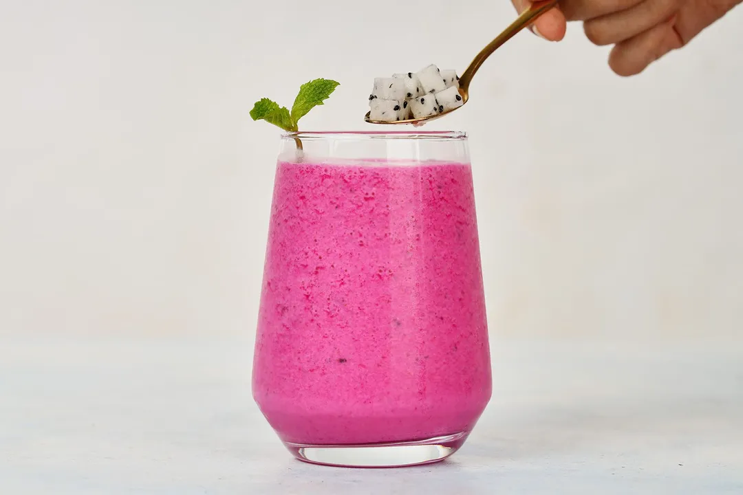 A hand holding a spoon filled with white dragon fruit hovering over a glass of pink Dragon Fruit Smoothie.