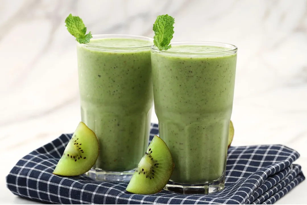 Two glasses of kiwi smoothie placed on a blue cloth with slices of green kiwi leaning onto their sides.