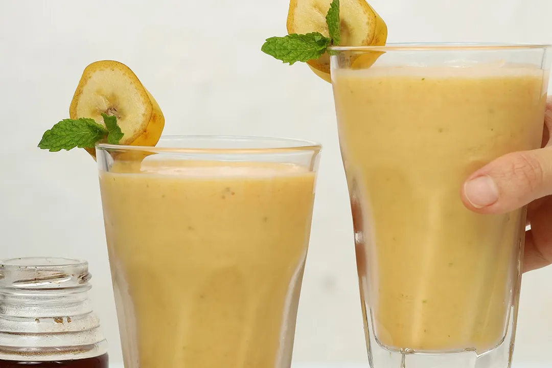 A hand picking up a glass of Mango Banana Smoothie placed next to an identical glass.