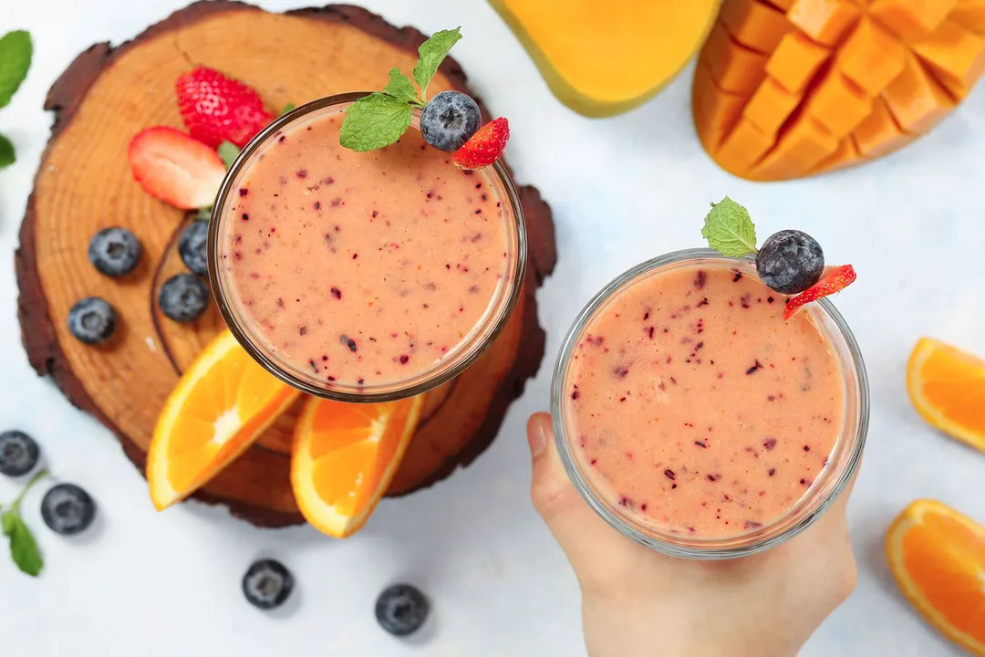 A high-angle shot of two glasses of Mango Berry Smoothie surrounded by blueberries, strawberries, orange wedges, and scored mangoes.