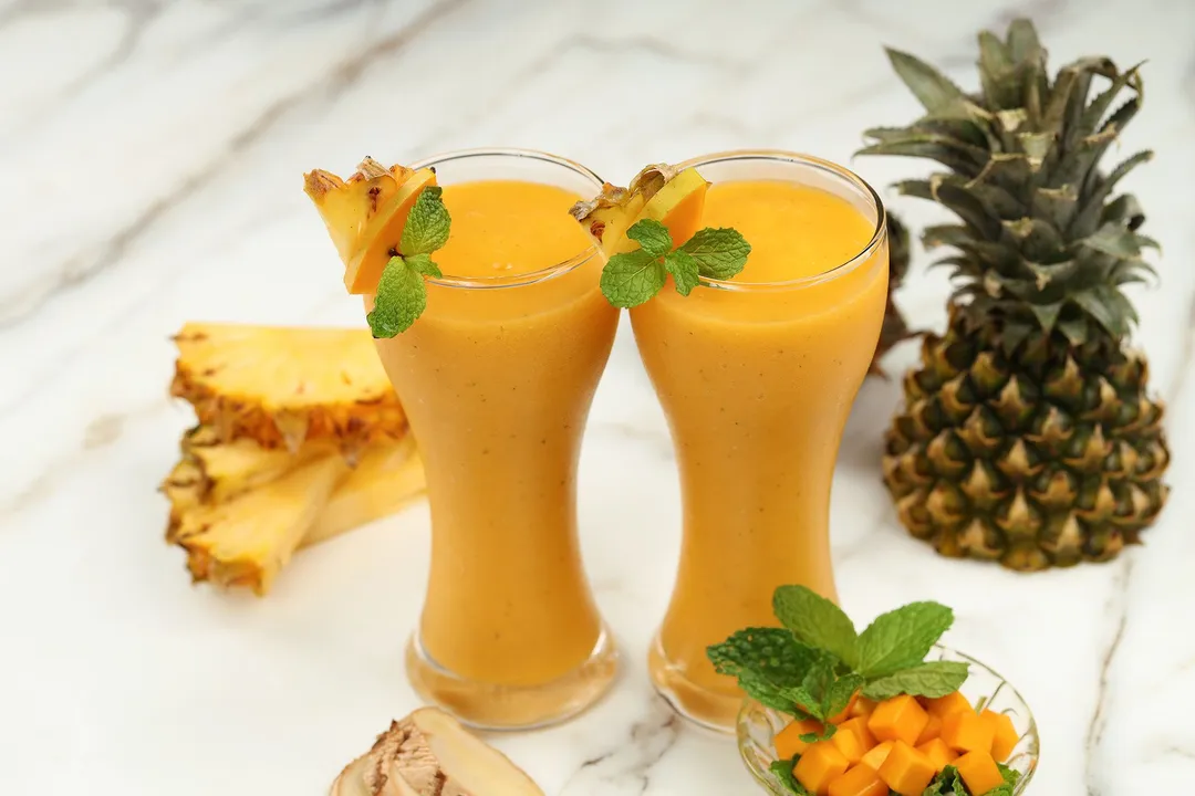 Two tall glasses of Mango Ginger Smoothie placed near pineapple slices, sliced mangoes, and ginger.