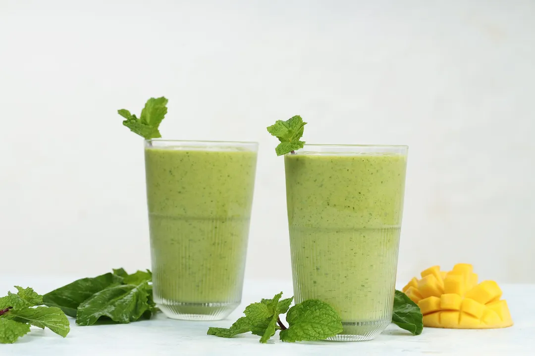 Two glasses of Mango Spinach Smoothie placed near fresh spinach, mint leaves, and a scored mango.