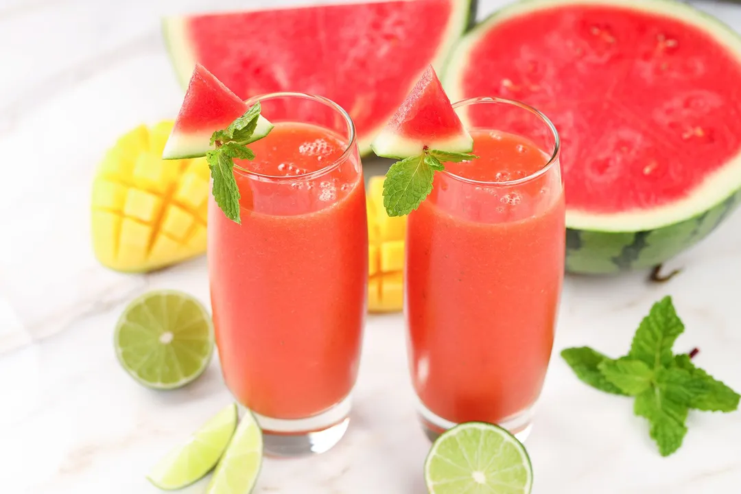 Two glasses of Mango Watermelon Smoothie placed near lime wedges, sliced watermelon, mint leaves, and scored mangoes.