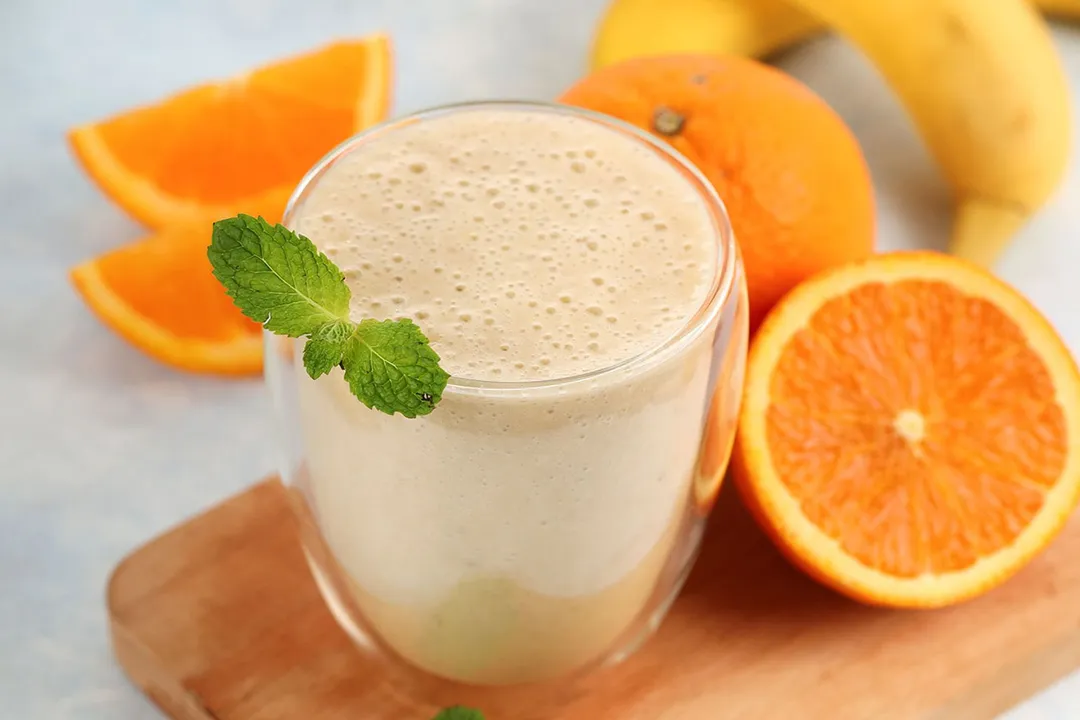 A small glass of Orange Banana Smoothie placed on a wooden board near orange slices.