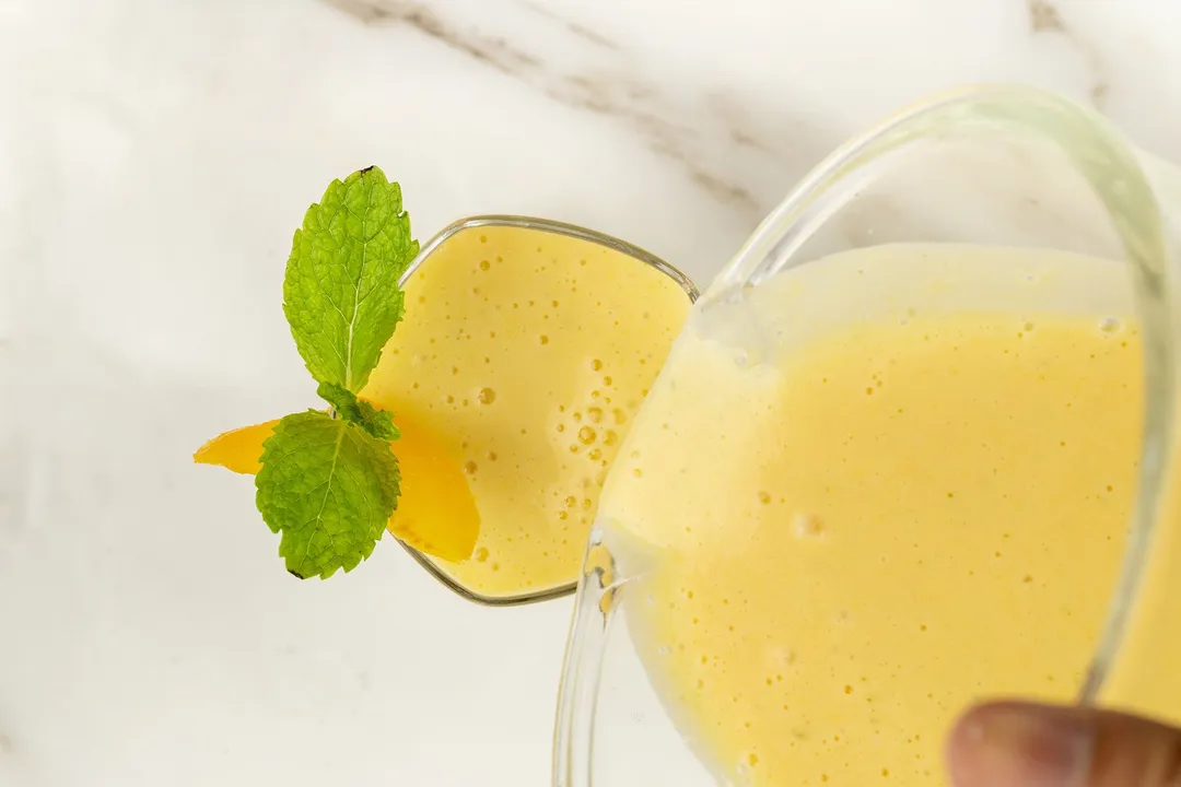 A high-angle shot of a blender pouring a peach smoothie into a glass garnished with a mint leaf.